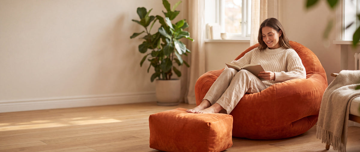 Femme lisant un livre, installée dans le fauteuil pouf Le Tone orange en velours côtelé, pieds sur son repose-pieds