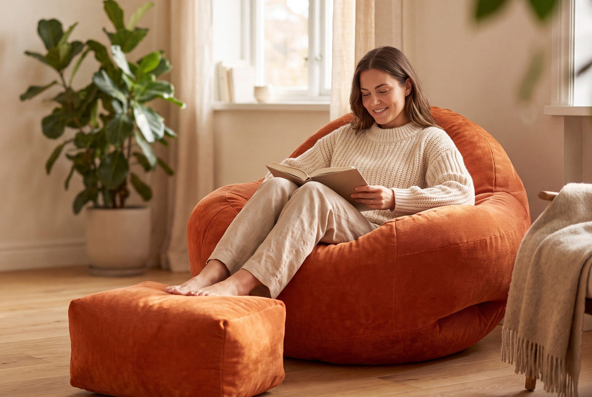 Femme lisant un livre, installée dans le fauteuil pouf Le Tone orange en velours côtelé, pieds sur son repose-pieds
